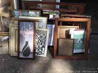 Group of framed pictures and empty frames placed outdoors on pavement showing variety of artworks and frame styles.