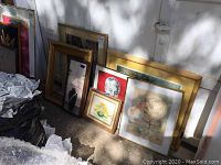 General view of six framed pictures and one empty frame leaning against wall outdoors showing wear and sunlit shading.
