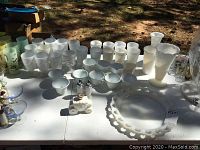 Wide view of milk glass collection showing cups, glasses, salt and pepper shakers, and vases arranged on table outdoors.