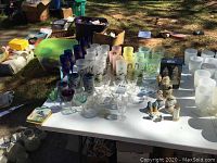 Overview of table displaying assorted glassware and salt and pepper shakers outdoors including collectible glasses, champagne flutes, and various drinking glasses.
