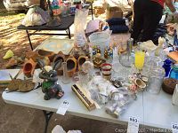 Table display of assorted small decorative items including wooden clogs, glasses, napkin rings, and figurines on a white folding table outdoors