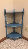 Blue wooden three-tier corner shelf against beige wall and carpet floor, showing front and side angles with turned spindle supports and decorative finials.