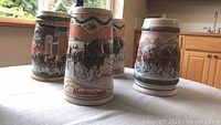 Four Budweiser beer steins on a white tablecloth showing detailed colored scenes with horses and landscapes