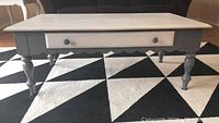 Front view of wooden coffee table showing white top and gray painted legs with drawer and round knobs.