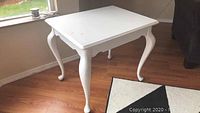 Front and side view of square white wooden accent table with cabriole legs on wood floor near window and black furniture.