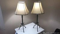 Pair of table lamps on white surface under warm light, showing full view of both lamps with beige fabric shades and metal tripod bases.