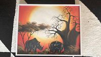 Framed colorful African art painting depicting two rhinoceroses, two birds, a large prominent tree on the right, smaller tree on the left, against a vivid sunset background.