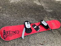Red snowboard with black graphic character designs and black/silver bindings viewed from above on a pavement surface.