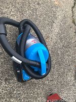 Top angled view of the vacuum cleaner showing the blue top, clear dust container, and black flexible hose coiled around the cleaner on concrete ground.
