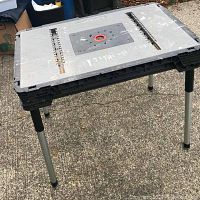 Folding utility table set up outdoors on a concrete surface, showing the top work surface with slots and red detail in middle.