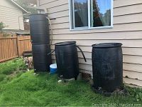 Wide view of four large black rain water barrels outside a house, showing barrels stacked and side by side with connected hoses
