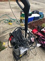 Power washer front with handle and coiled hose placed on driveway with other gardening items.