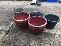 Seven plastic planters of different colors and sizes positioned on pavement. Two large red ones, two dark green ones, one grey one, and three small nested black shallow planters visible.