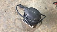 Black electric handheld finishing sander with power cord wrapped around it on concrete floor.