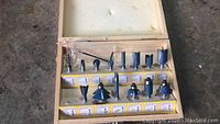 Top view of the open wooden case containing 16 blue and black router bits with foam padding inside the lid. Various shapes and sizes, some with bearing guides.