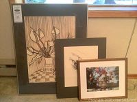 Three framed artworks arranged leaning against a window: largest with lily flower line art, medium with bird drawing, and smallest colorful floral print