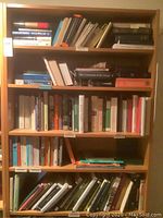 Front view of a tall wooden bookcase filled with books on five shelves. Light wood, dimensions approx 35.5 x 11 x 79.5 inches.