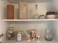 Overview of the lot with jugs, vase, decanter, and cups in white shelving unit