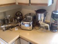 Photo showing several kitchen appliances on a countertop including a shiny stainless steel 4-slice toaster, coffee urn with glass carafe, manual metal meat grinder, metal kettle, large metal stock pot with lid, strainer, and Salad Maid spinner.