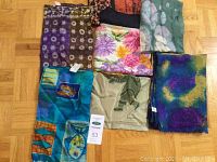 Seven scarves spread out on wooden floor, showing different colors and patterns including floral, abstract and geometric designs in silk and wool materials.