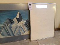 Two framed artworks leaning against wall, showing the stylized mountain painting and a white framed piece.