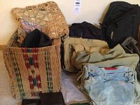 Photo showing various bags including a woven basket-style bag, fabric bags, and black backpacks arranged on the floor.