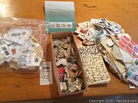 Wide view of all lot items spread on wooden table including bag of loose stamps, box with many small pieces of paper with stamps, and stack of paper with stamps attached.
