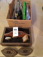 Photo showing a cardboard box with metal shoehorns, brushes, and other tools behind a wooden box containing shoe polish tins, brushes, and two vintage irons in front.