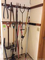 Wall of garden tools hanging on wooden racks, showing multiple garden implements with wooden and metal parts.