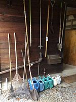 Photo of long-handled garden tools arranged against a wooden wall with plastic watering cans on the ground in front, showing a variety of spades, rakes, pitch fork, and watering cans.