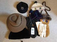 Overview of all hats and gloves arranged on carpet including a beige wide-brimmed hat, black cap, white knit hat, Inuit mittens, beige gloves, black mittens, and black fur hat.