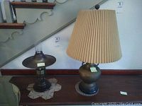 Two metal table lamps displayed on a wooden surface with a staircase in the background. One lamp has a wide metal shade with clear glass chimney, the other has a pleated fabric lampshade.