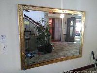 Full view of large rectangular mirror with decorative gold frame hanging on a wall, reflecting plants, stairs and furniture in room.
