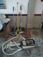 Photo showing cleaning tools including mop, brooms, Endust sweeper, vintage shoeshine box, and Eureka vacuum hose attachments on carpet