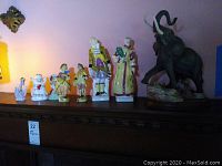 Group photo of seven figurines including couple, angels, elephant and small figures on wooden shelf.