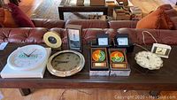 Assortment of clocks laid out on a wooden table showing variety in shapes, sizes, and brands.