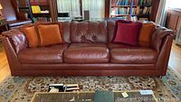 Front view of brown leather sofa with four throw pillows (two orange, one burgundy). Sofa has tufted back cushions and seat cushions.