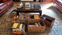 Wide view of the collection showing stacks of hardcover and paperback books on a coffee table and in boxes on a carpeted living room floor.