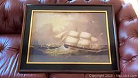 Framed nautical wall art on a brown leather surface, showing a detailed sailing ship with three sails in stormy seas.