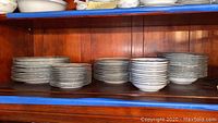 Five stacks of dinnerware on a wooden shelf including plates of various sizes and bowls, all white with a blue-gray floral and garland border design.