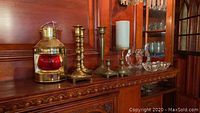 Wide view of brass lantern, two brass candlesticks, glass candle holders, and small glass dishes on wooden shelf