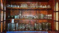 Wide view of entire glassware collection arranged on wooden shelves showing a variety of glass styles and sizes including pilsners, shot glasses, and more.