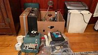 Photo showing various office supplies including an Olivetti adding machine, briefcases, a shredder, and boxes with office items on wooden floor.
