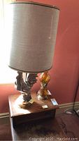 Photo of two lamps on a wooden side table; left lamp has a ceramic rooster base with beige lampshade; right lamp is amber-colored glass.