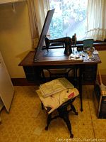 Wide view showing Singer treadle sewing machine with wooden top and drawers, accompanying wooden stool, fabric cover, scissors, and other sewing accessories on the table.