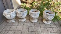 Front view of four cement planters on patio, showing Greek key pattern, ornamental face details, soil inside, some chips and nicks.
