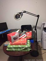 Photo of table with stacked reams of copy paper in green and orange packaging, various office supplies, and black floor lamp with magnifier on adjustable arm.