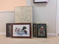 Wide view showing four framed pieces grouped on floor leaning against wall, includes two artworks of bird and child, and two maps.