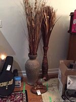 Two tall decorative vases placed on floor; one ceramic vase and one banana leaf wrapped vase, both with decorative sticks inside.