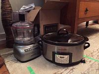 Side view of KitchenAid food processor next to Crock-Pot slow cooker on carpeted floor, with the food processor bowl and lid on top and the slow cooker lid also visible.